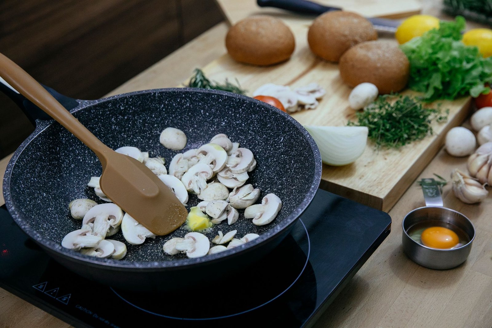 From above of silicone spatula in pan with frying chopped mushrooms placed on electric stove near wooden cutting board with assorted herbs and vegetables in kitchen